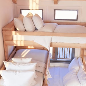 May include: A wooden bunk bed with white bedding and pillows. The top bunk has a white duvet and three pillows, while the bottom bunk has a white duvet and four pillows. The bed is in a room with a window and natural light.