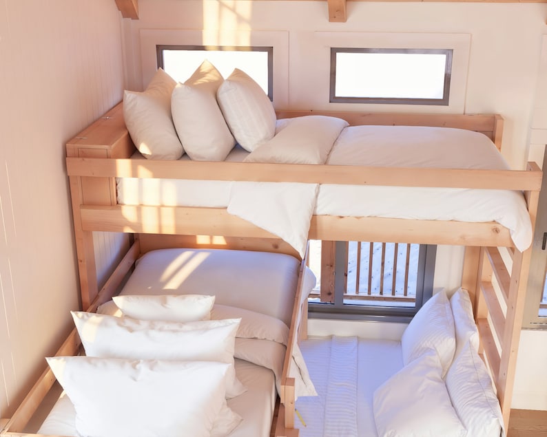 May include: A wooden bunk bed with white bedding and pillows. The top bunk has a white duvet and three pillows, while the bottom bunk has a white duvet and four pillows. The bed is in a room with a window and natural light.