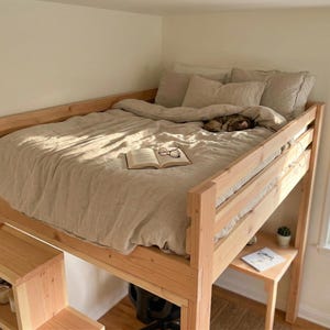 May include: A wooden loft bed with a light-colored comforter and pillows. An open book and glasses rest on the bed. A small cat is sleeping under the covers. A small desk and bookshelf are attached to the bed.