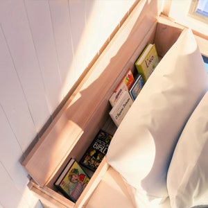 May include: A wooden bed frame with a built-in shelf for storing books. The shelf is filled with a variety of books, including a book titled "Lino" and another titled "O Segredo do Anjo". The bed is made with white bedding and two pillows.