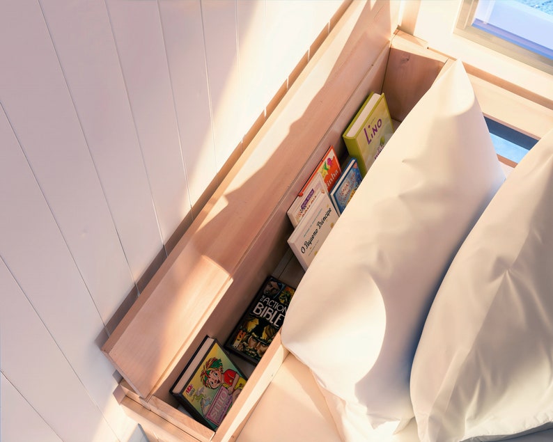 May include: A wooden bed frame with a built-in shelf for storing books. The shelf is filled with a variety of books, including a book titled "Lino" and another titled "O Segredo do Anjo". The bed is made with white bedding and two pillows.