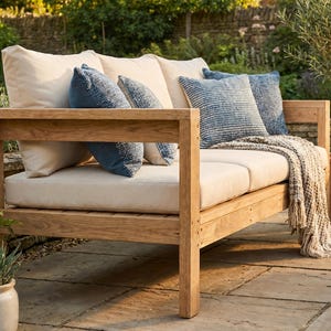 Puede incluir: Un sofá de exterior de madera con cojines color crema y varias almohadas azules estampadas. Una manta de tonos neutros está sobre el sofá. El sofá está colocado en un patio de piedra, con plantas en macetas en el fondo.