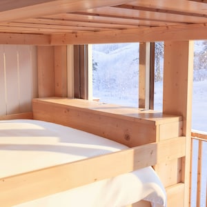 May include: A wooden bunk bed with a white sheet on the bottom bunk. The bed is positioned in front of a window with a view of a snowy landscape.