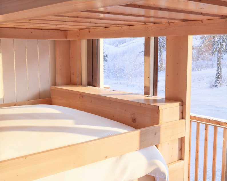 May include: A wooden bunk bed with a white sheet on the bottom bunk. The bed is positioned in front of a window with a view of a snowy landscape.