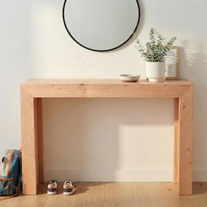 Puede incluir: Una consola de madera de color claro con una parte superior rectangular y patas gruesas. Un espejo redondo negro cuelga de la pared encima de la mesa. Una maceta blanca pequeña con vegetación, un cuenco pequeño y una imagen enmarcada están sobre la mesa. Una mochila y zapatos están en el suelo.