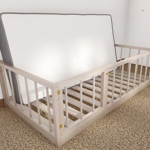May include: A white wooden toddler bed rail with a gray mattress inside. The bed rail has a gate on one side that can be opened and closed.