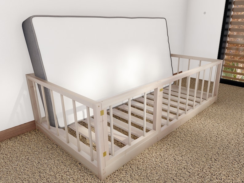 May include: A white wooden toddler bed rail with a gray mattress inside. The bed rail has a gate on one side that can be opened and closed.