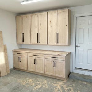 May include: Light wood cabinets and drawers with black hardware are mounted on a wall. The cabinets are above a countertop and a white door is to the right. Several pieces of wood are leaning against the wall.