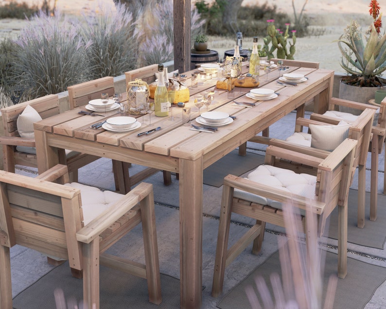 Op de afbeelding: Een houten eettafelset voor buiten met een lange rechthoekige tafel en zes stoelen. De tafel heeft een natuurlijke houten afwerking en de stoelen hebben witte kussens. De set is op een terras met een stenen vloer en omringd door planten geplaatst.