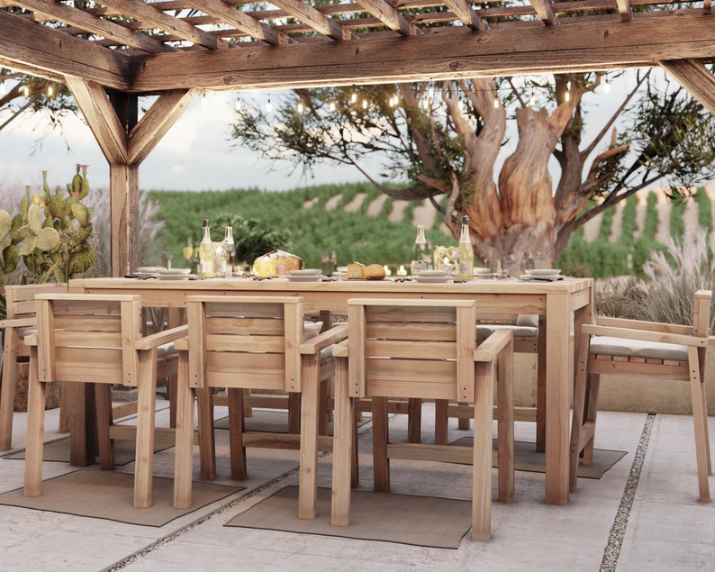 Op de afbeelding: Een houten eettafelset voor buiten met een rechthoekige tafel en zes stoelen. De tafel en stoelen zijn gemaakt van natuurlijk hout en hebben een rustieke afwerking. De set staat onder een houten pergola met lichtslingers. De achtergrond toont een wijngaard met een grote boom in het midden.