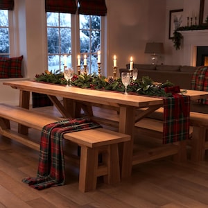 May include: A wooden dining table with two benches, set for a holiday meal. The table is decorated with a garland, candles, and plaid runners. The room has large windows and a fireplace.