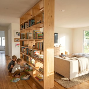 Może przedstawiać: Jasna drewniana półka na książki wypełniona książkami, dzieląca przestrzeń mieszkalną. Dwoje dzieci ogląda książki na podłodze w pobliżu półki. Pokój ma podłogi z twardego drewna i sofę w neutralnych odcieniach.