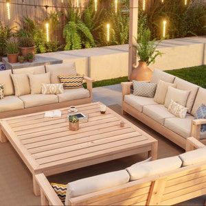 May include: A wooden outdoor patio furniture set with a large square table and three couches. The couches have beige cushions and are made of natural wood. The table is also made of natural wood and has a slatted top. The furniture is set on a tan rug and there are string lights hanging overhead.