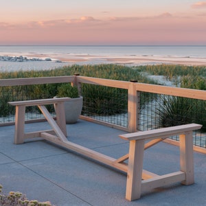 May include: A wooden outdoor table with a long, rectangular top and a simple, rustic design. The table is made of unfinished wood and has a natural, light brown colour. The table is set against a backdrop of a sandy beach and a blue sky with pink clouds.
