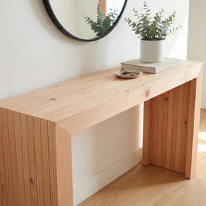 Może przedstawiać: Jasny drewniany stolik konsolowy z prostokątnym blatem i pionowym lamelowym wzorem po bokach. Nad stołem wisi okrągłe czarne lustro. Na stole stoi mała roślina w białej doniczce.