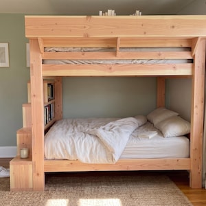 May include: A light-colored natural wood bunk bed featuring a built-in staircase with integrated shelving on the side. The lower bunk is dressed with white bedding and pillows, while the upper bunk has a wooden canopy frame and a mattress.