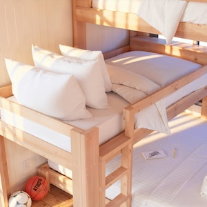 May include: A wooden bunk bed with white bedding and pillows. The top bunk has a white sheet and a white pillow. The bottom bunk has a white sheet, a white pillow, and a white blanket. There is a basketball on the floor next to the bed.