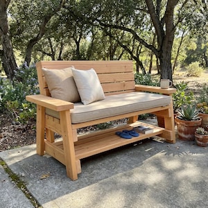 May include: A wooden outdoor bench with a light brown finish, featuring a slatted back and armrests. It has a beige cushion and two pillows. A lower shelf holds books and blue sandals. A mug sits on the armrest. Potted plants are nearby.