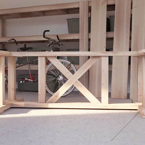 May include: A wooden table base with an X-shaped support structure. The base is made of unfinished wood and has a natural colour. The base is sitting on a concrete floor.