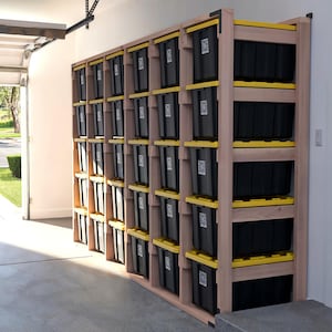 May include: A wooden storage unit with multiple shelves filled with black storage bins with yellow accents. The unit is positioned inside a garage, offering a practical storage solution. The garage door is partially open, revealing the outside.