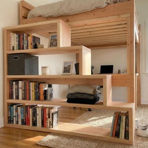 May include: A light-colored wooden loft bed with built-in shelving and a desk area. The shelves are filled with books, a storage box, and decorative items. A laptop sits on the desk. The bed is made of light-colored wood.