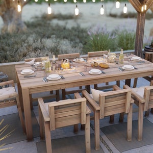 Op de afbeelding: Een houten eettafel voor buiten met zes stoelen, gedekt voor een maaltijd met borden, bestek en glazen. De tafel is gemaakt van natuurlijk hout en heeft een rustieke uitstraling. De stoelen zijn ook van hout en hebben kussens.
