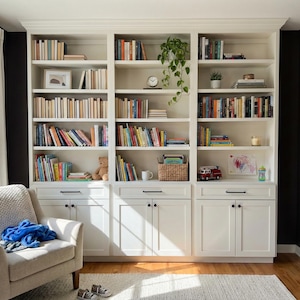 Könnte beinhalten: Ein weißer Einbaubuchschrank mit mehreren Regalen, die mit Büchern verschiedener Größen und Farben gefüllt sind. Der Bücherschrank hat untere Schränke mit schwarzen Griffen. Vor dem Bücherschrank steht ein beiger Sessel mit einer blauen Decke.