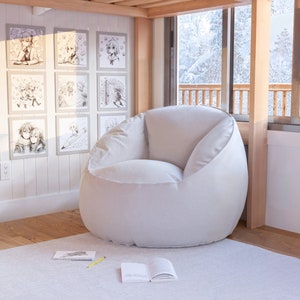 Puede incluir: Un sill&oacute;n de puf blanco en una habitaci&oacute;n con suelo de madera y una ventana con vistas a un bosque nevado. Hay dibujos enmarcados en la pared detr&aacute;s del sill&oacute;n.