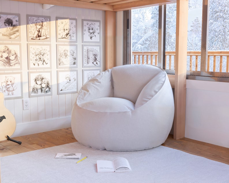 Puede incluir: Un sill&oacute;n de puf blanco en una habitaci&oacute;n con suelo de madera y una ventana con vistas a un bosque nevado. Hay dibujos enmarcados en la pared detr&aacute;s del sill&oacute;n.