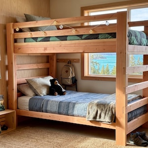 Op de afbeelding: Een lichtbruin houten stapelbed met een ladder. Het bovenste bed heeft een groen en zwart patroon dekbed, terwijl het onderste bed een blauw en wit gestreept dekbed en een beige deken heeft. Een zwarte teddybeer zit op het onderste bed. Het bed staat in een kamer met een raam.