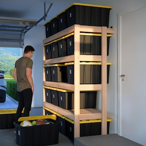May include: A wooden storage shelf unit with multiple black storage bins with yellow lids. The shelf unit is in a garage with a white door and a car in the background.