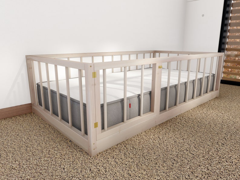 May include: A white wooden crib with a gray mattress and a white wooden gate. The crib is in a room with a carpeted floor.