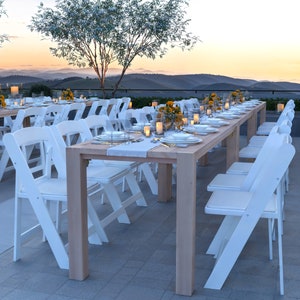 Op de afbeelding: Een lange houten tafel met witte klapstoelen, gedekt voor een diner. De tafel is versierd met kaarsen en bloemen. De tafel is gedekt met witte borden, bestek en glazen. De tafel staat op een terras met uitzicht op een bergketen in de verte.