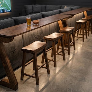 May include: A custom-built, dark wood bar with a Z-shaped leg design, illuminated by under-bar lighting. The bar is paired with several wooden stools. A glass of beer and a bowl of snacks sit on the bar. The bar is set up in front of a dark grey couch.