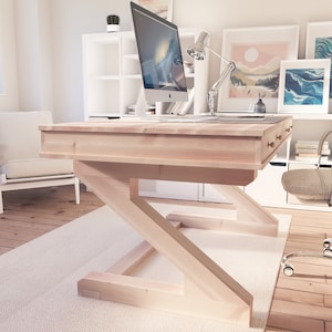 Puede incluir: Un escritorio de madera clara con una base en forma de Z y un caj&oacute;n. El escritorio est&aacute; en una oficina en casa con una silla blanca, un monitor de computadora y una l&aacute;mpara.