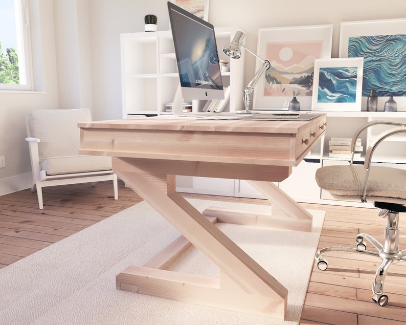 Puede incluir: Un escritorio de madera clara con una base en forma de Z y un caj&oacute;n. El escritorio est&aacute; en una oficina en casa con una silla blanca, un monitor de computadora y una l&aacute;mpara.