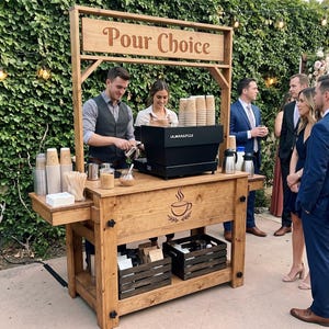 May include: A wooden mobile coffee cart featuring a sign that reads Pour Choice, a black espresso machine, stacks of paper cups, wooden stirrers, and two dark wooden crates on the lower shelf. The front panel displays a graphic of a coffee cup.
