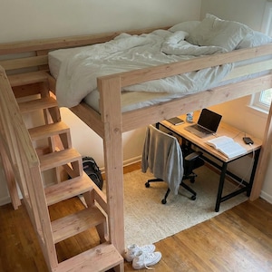 May include: A wooden loft bed with a built-in staircase. The bed frame is natural wood, and the bed is made with a white comforter. A desk with a laptop and chair is underneath the bed. The room has hardwood floors.