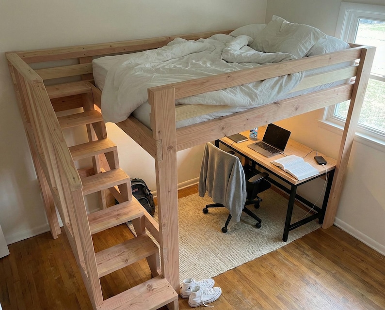 May include: A wooden loft bed with a built-in staircase. The bed frame is natural wood, and the bed is made with a white comforter. A desk with a laptop and chair is underneath the bed. The room has hardwood floors.
