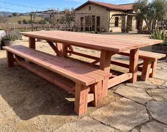 May include: A large rectangular wooden dining table with a natural reddish-brown finish, featuring thick plank construction and sturdy legs with cross-bracing support, accompanied by two matching long benches of the same wood and design style.