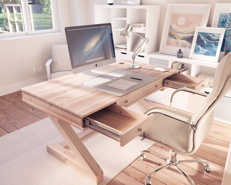 Puede incluir: Un escritorio de madera clara con un monitor de computadora blanco, una silla blanca y una unidad de estanter&iacute;a blanca en el fondo. El escritorio tiene dos cajones que est&aacute;n abiertos, revelando el interior de madera. El escritorio tiene un dise&ntilde;o simple y moderno.