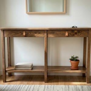 May include: A wooden console table with two drawers and a lower shelf. The table is made of stained wood with leather drawer pulls. A small potted plant and books are on the shelves. A mirror hangs above the table.