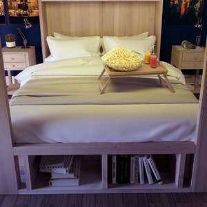 May include: A wooden bed frame with a white bedspread and pillows. A wooden tray with a bowl of popcorn and two glasses sits on the bed. The bed has built-in shelves underneath for storage.