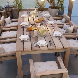 Op de afbeelding: Een houten eettafel voor buiten met zes stoelen, gedekt voor een maaltijd met borden, bestek en glazen. De tafel is gemaakt van natuurlijk hout met een rustieke afwerking. De stoelen zijn ook van hout en hebben kussens.