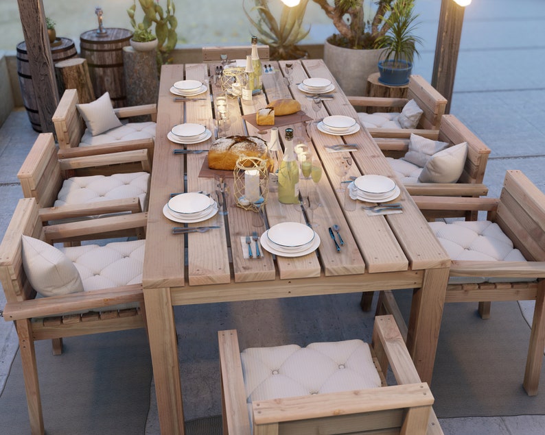 Op de afbeelding: Een houten eettafel voor buiten met zes stoelen, gedekt voor een maaltijd met borden, bestek en glazen. De tafel is gemaakt van natuurlijk hout met een rustieke afwerking. De stoelen zijn ook van hout en hebben kussens.