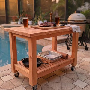 Puede incluir: Un carrito de parrilla de madera para exteriores con un estante inferior y ruedas. El carrito contiene una tabla de cortar con carne en rodajas, dos vasos de cerveza y una sartén de hierro fundido. Una parrilla ahumadora está en el fondo.