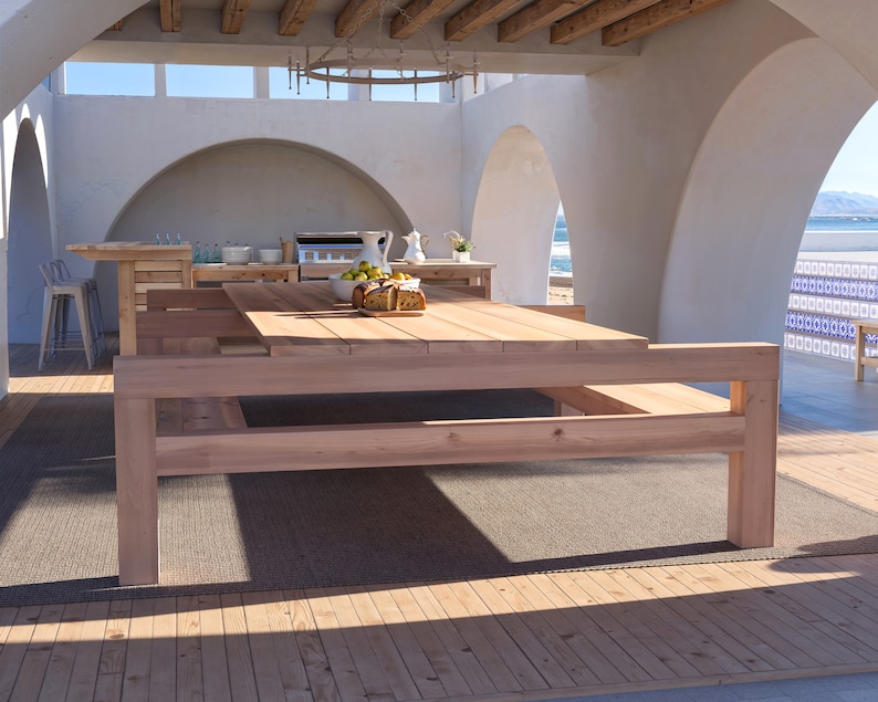 K&ouml;nnte beinhalten: Ein gro&szlig;er Holztisch mit zwei B&auml;nken auf jeder Seite, aufgestellt auf einer Terrasse mit Blick auf das Meer. Der Tisch ist aus Naturholz gefertigt und hat einen rustikalen Look. Die B&auml;nke sind ebenfalls aus Holz gefertigt und haben ein schlichtes Design.