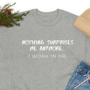Peut inclure: Un sweat-shirt gris avec le texte "NOTHING SURPRISES ME ANYMORE... I WORK IN HR." en lettres blanches. Le sweat-shirt est présenté sur un fond blanc avec une paire de chaussures noires, un jean bleu et un bonnet beige.