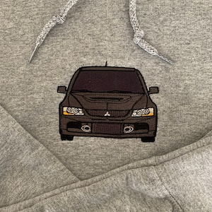 Może przedstawiać: Szara bluza z haftowanym czarnym obrazem samochodu z przodu. Samochód ma czarny grill i reflektory oraz czarny zderzak z dwoma rurami wydechowymi.
