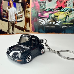 May include: Black Porsche 911 keychain with a silver chain and ring. The keychain is in focus, and the background is a blurry image of a movie poster for "The Fast and the Furious: Tokyo Drift".